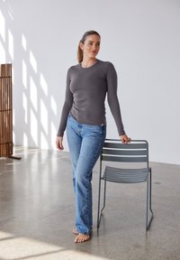Grå långärmad topp i kombination med ljusblå jeans, står barfota bredvid en grå metallstol. Slät textur, åtsittande design.