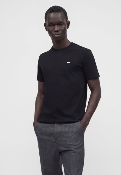 Homme à la peau foncée portant un t-shirt noir à manches courtes et un pantalon gris à fines rayures, debout les mains dans les poches devant un fond uni.