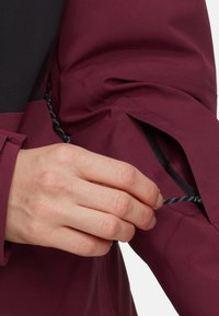 Mörkröd jacka med vattentåligt tyg, utrustad med en ficka med dragkedja och en justerbar snodd. Närbild av en hand som greppar snodden.