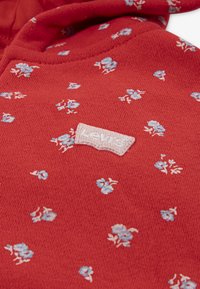Textile rouge avec des motifs floraux éparpillés en bleu et blanc, présentant un patch logo blanc en relief.