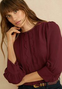 Tamsiai raudona palaidinė su pūstomis rankovėmis, bangelėmis apdaila ir vertikaliomis klostėmis. Yra mygtuko akcentai ir apvalus kaklo iškirpimas. Tekstūruota medžiaga.