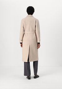 Cappotto in lana beige con vestibilità su misura, dotato di uno spacco sul retro, vita cintata e polsini abbottonati. Abbinato a pantaloni larghi neri.
