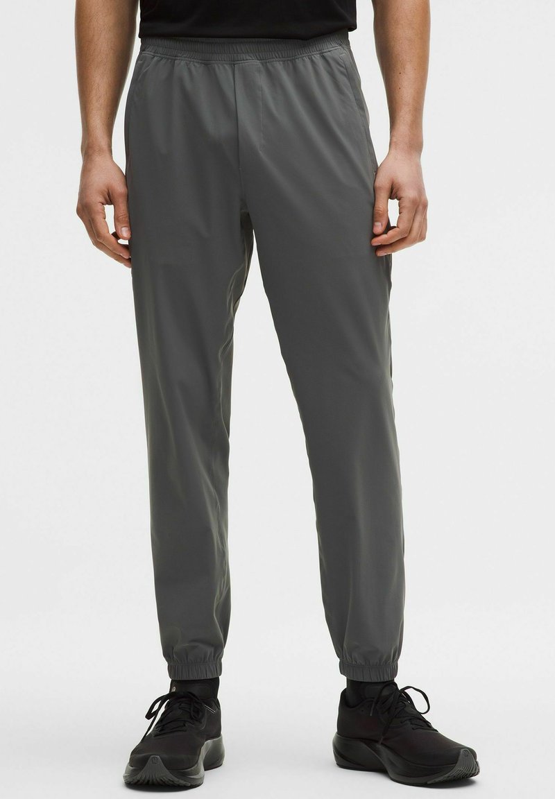 Uomo che indossa pantaloni jogger grigio scuro con vita elastica e sneakers nere, in piedi su uno sfondo chiaro e uniforme.