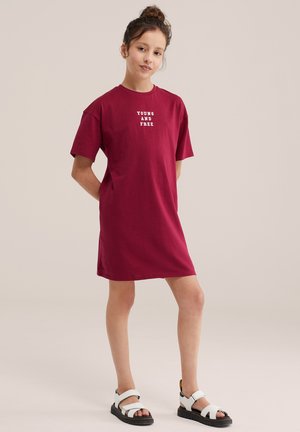 Jong persoon staand, gekleed in een kastanjebruine t-shirtjurk met de tekst "YOUNG AND FREE" en witte sandalen met zwarte zolen.