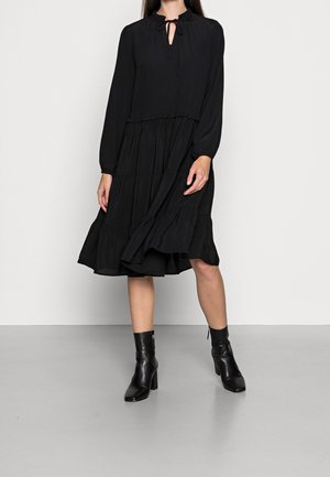 Schwarzes schichtweises Kleid mit langen Puffärmeln, Schlüsselloch-Ausschnitt und Rüschensaum. Kombiniert mit schwarzen Knöchelstiefeln. Glatte Stofftextur.