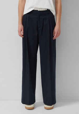 Personne portant un pantalon large bleu marine, un haut blanc rentré et des baskets blanches à semelles marron, debout contre un mur gris uni.