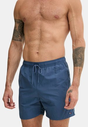 Uomo senza maglietta che indossa pantaloncini da bagno blu con coulisse e tasche laterali, mostrando tatuaggi su entrambe le braccia e un tatuaggio sul polso della mano destra.