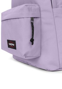 Zaino viola con una tasca frontale, chiusura a zip e cordino nero. Presenta un fondo arrotondato e una patch con il logo Eastpak. Texture liscia.