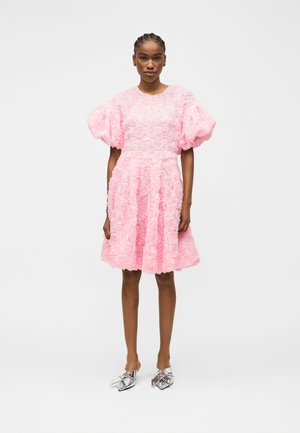 Femme portant une robe rose mi-longue avec des manches bouffantes et une texture florale, associée à des chaussures argentées à bout pointu, debout devant un fond uni.