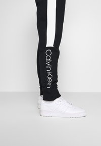 Nohy v černých teplákách Calvin Klein s bílým pruhm na straně a bílých teniskách Adidas na světle šedé podlaze a pozadí.
