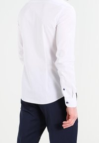 Chemise blanche à manches longues avec poignets boutonnés, dotée d'un col classique et d'une texture lisse, portée avec un pantalon foncé.