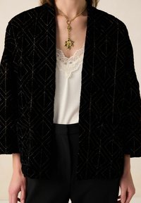 Zwarte fluwelen jas met geometrisch patroon en reflecterende accenten, open voorkant, gecombineerd met een wit kanten topje en zwarte broek. Gouden kettingdetail.