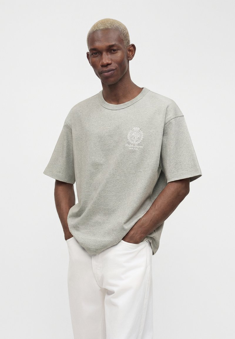 Homme aux cheveux blonds courts portant un T-shirt Ralph Lauren gris clair et un pantalon blanc, debout les mains dans les poches devant un fond uni.