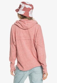 Sudadera rosa jaspeada con un ajuste holgado, que cuenta con capucha y mangas largas. Combinada con un sombrero tipo pescador rosa con estampado floral. Pantalones vaqueros claros.