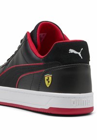 Sneaker in pelle nera con accenti rossi, dotato di una suola in gomma bianca, retro testurizzato e logo giallo Ferrari sul lato.