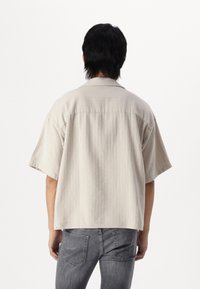 Persona con capelli neri corti che indossa una camicia beige a maniche corte e larga, e jeans grigi sbiaditi, di spalle su uno sfondo bianco semplice.