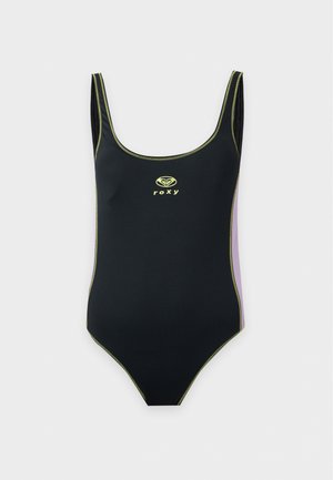 Une femme présente un maillot de bain une pièce noir Roxy avec des bordures contrastantes sur un fond clair et uni.