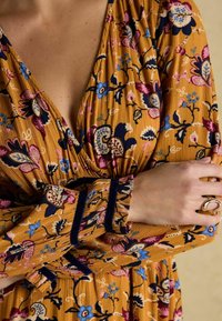 Senfgelbes Kleid aus Stoff mit floralen Mustern in Blau, Pink und Schwarz. Verfügt über einen V-Ausschnitt und umgeschlagene Ärmel mit dunklem Besatz. Goldener Ring an der Hand.