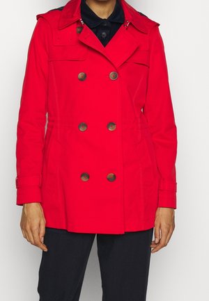 Persoon die een felrood tweeregelig trenchcoat draagt met zes bruine knopen en een zwarte broek, staand tegen een lichte achtergrond.