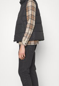 Chaleco acolchado negro con costuras horizontales, llevado sobre una camisa de cuadros en beige y marrón. El modelo también lleva vaqueros oscuros, de pie de lado.