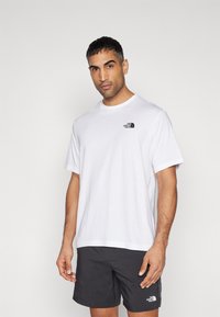 Camiseta de algodón blanca con cuello redondo y mangas cortas, que presenta un logo negro en el lado izquierdo del pecho. Textura de tejido suave.