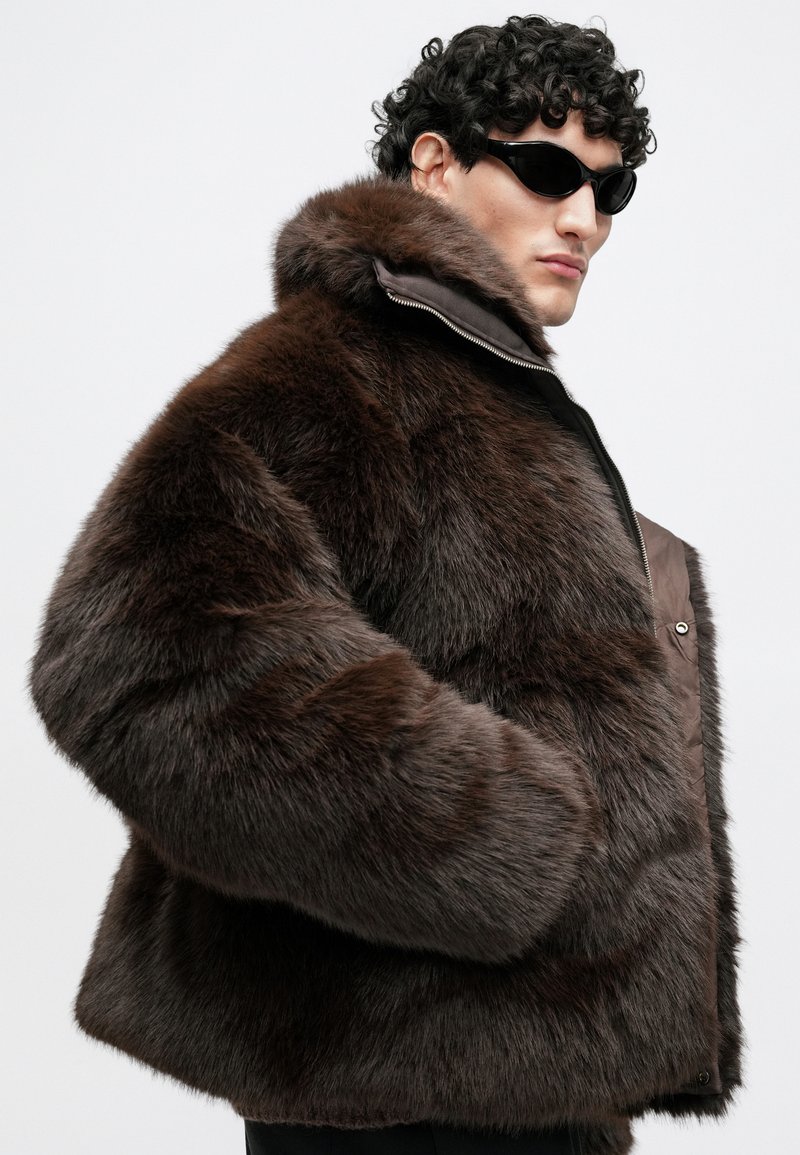 Brown faux fur jacket with a wide collar, zip fastening, and side pockets. Features a smooth leather panel and glossy texture.