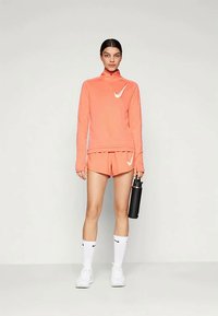 Nike sportinė apranga koralinės spalvos, sudaryta iš užtrauktuko ilgarankovių marškinėlių ir šortų. Papuošta baltais logotipais ir derinama su juodu vandens buteliuku.