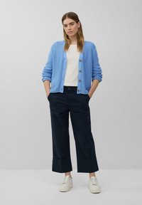 Helles blaues Strickjacke mit Knöpfen, kombiniert mit einem weißen Oberteil und navyblauen weit geschnittenen Hosen. Weiße Sneakers runden das Outfit ab.