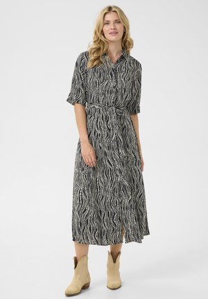 Black and white, short-sleeve midi dress with a wavy pattern, belted waist, and front slit. Model wears tan ankle boots.
