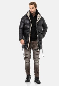 Schwarze Puffjacke mit beigem Futter, ausgestattet mit einer Kapuze und zwei Fronttaschen, kombiniert mit verwaschenen braunen Jeans und schwarzen Stiefeln.