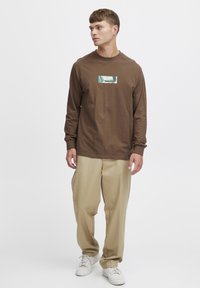 Long-sleeve brown cotton T-shirt with a rectangular graphic design in blue and green at the center, paired with light beige trousers.