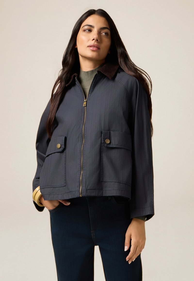 Veste cropped bleu marine avec fermeture éclair à l'avant, deux poches avant et col en velours côtelé brun. Texture lisse, design décontracté.