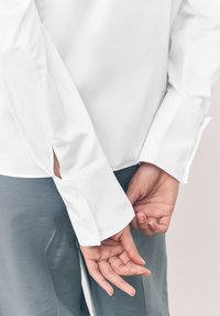 Chemise blanche à boutons avec manches longues, présentant des fentes latérales aux poignets. Tissu gris visible en dessous. Mains croisées dans le dos.