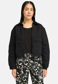 Black cropped jacket with a collared neck, featuring two front pockets and a zip closure, paired with floral-patterned black pants.