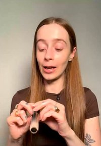 Junge Frau mit langen Haaren hält eine kleine Kosmetiktube und öffnet sie, während sie vor einem schlichten neutralen Hintergrund spricht.