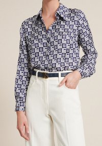 Navy blue patterned shirt with geometric white designs, long sleeves, button-up front, paired with white trousers and a dark belt.