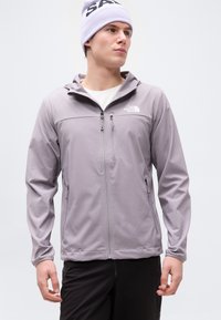 Άνδρας που φοράει ανοιχτό μοβ ζακέτα με φερμουάρ και το λογότυπο The North Face, μαύρο παντελόνι και ανοιχτό μοβ σκουφάκι, κοιτάζοντας προς τα πλάγια μπροστά σε απλό φόντο.