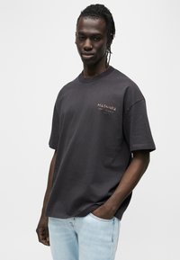 Σκούρο γκρι βαμβακερό μπλουζάκι με κοντά μανίκια και στρογγυλή λαιμόκοψη. Διαθέτει μικρό, ξεθωριασμένο λογότυπο σε αντίθετο χρώμα στο στήθος.