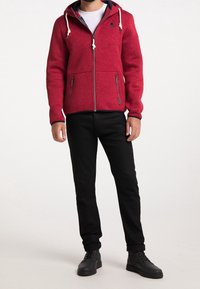 Punainen fleecepaita, jossa on vetoketjullinen huppari vuorattulla hupulla, nyörikiinnitykset ja kaksi vetoketjullista taskua. Yhdistetty mustiin housuihin ja mustiin kenkiin.