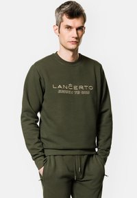 Oliwkowy sweter z dekoltem w serek, zdobiony wyraźnym złotym napisem "LANCERTO DRIVEN TO WIN" na piersi. Miękka tekstura, luźny krój.