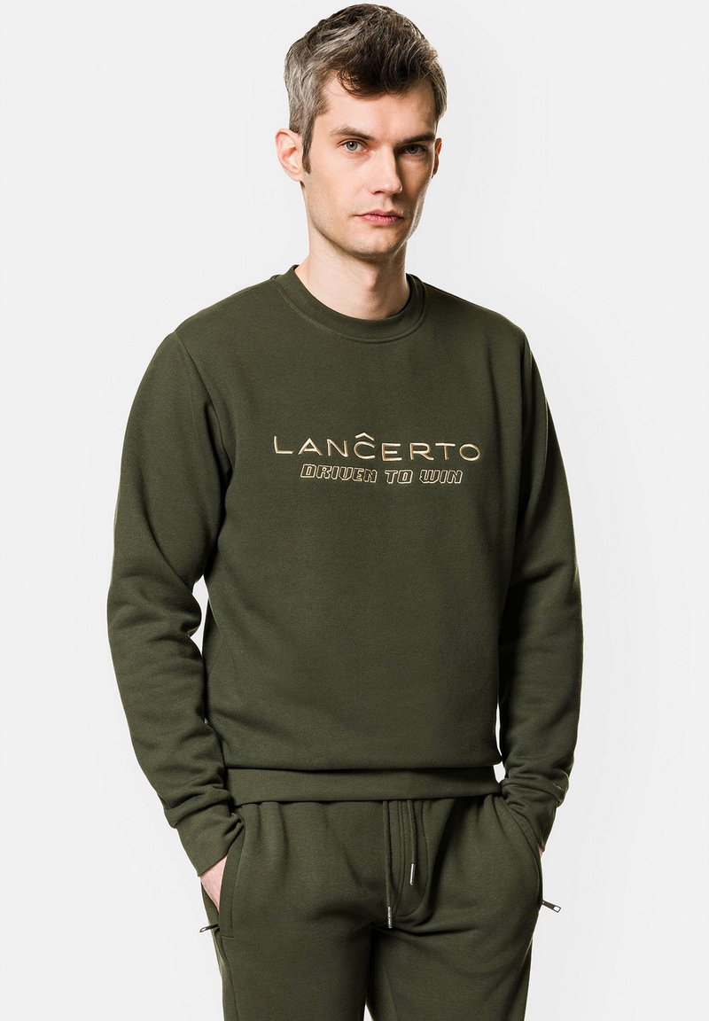 Oliwkowy sweter z dekoltem w serek, zdobiony wyraźnym złotym napisem "LANCERTO DRIVEN TO WIN" na piersi. Miękka tekstura, luźny krój.