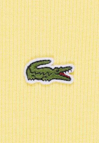 Gele gebreide stof heeft een groene geborduurde krokodillenlogo, omlijnd in het wit. De textuur is fijn en geribbeld.