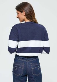 Pull rayé bleu marine et blanc avec un col côtelé et des boutons d'épaule, associé à un jean bleu foncé avec des coutures distinctives sur les poches arrière.