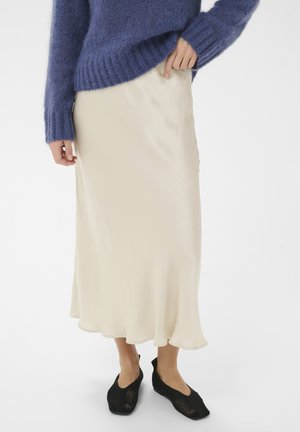 Osoba nosiaca krémovú saténovú midi sukňu s modrým chlpatým svetrom a čiernymi sieťkovanými teniskami na zapínanie, stojaca pred bielym pozadím.