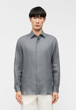 Man met een grijs, langarmig overhemd met knopen en witte broek, staand tegen een effen witte achtergrond.