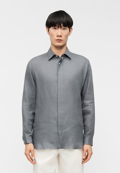 Homme portant une chemise grise à manches longues boutonnée et un pantalon blanc, debout devant un fond blanc uni.