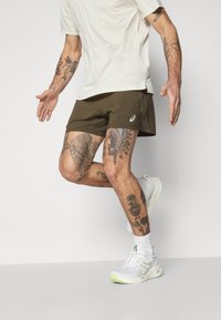 Men's lightweight, olive green athletic shorts with a slim fit, paired with white running shoes, showcasing intricate tattoos on the legs.