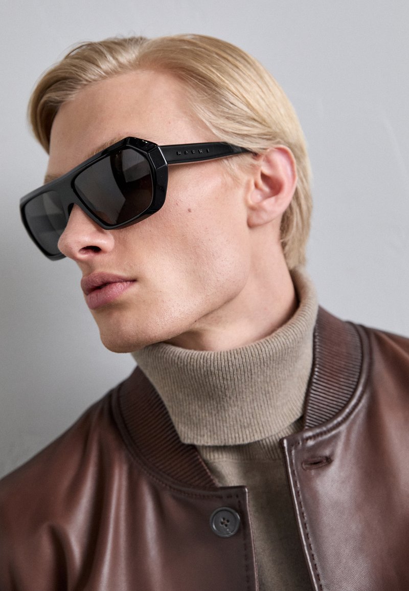 Mustat ylisuuret aurinkolasit kulmikkailla kehykset ja tummilla linsseillä, yhdistettynä beigeen poolopaitaan ja ruskeaan nahkabomber takkiin.