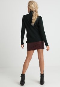 Dunkelgrüner Rollkragenpullover, taillierte Form, kombiniert mit einem burgunderfarbenen Leder-Minirock und schwarzen Stiefeletten mit klobigen Absätzen.
