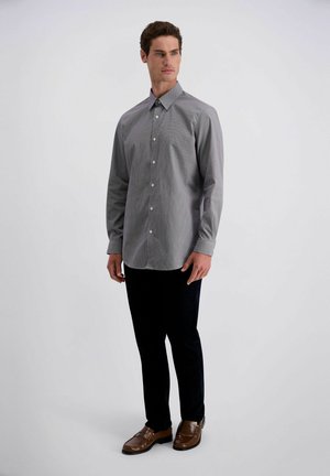 Man staat, draagt een grijs overhemd met knopen, zwarte broek en bruine nette schoenen tegen een effen lichte achtergrond.